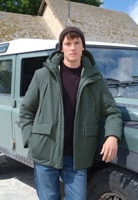 Grüne Steppjacke mit Kapuze, ausgestattet mit großen Fronttaschen und Reißverschluss. Über einem dunklen Pullover getragen, kombiniert mit blauen Jeans.