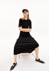 Schwarzes knielanges Kleid mit kurzen Ärmeln, geripptem Muster und lockerem Schnitt. Kombiniert mit spitzen schwarzen Ballerinas und einer schwarzen Mütze.