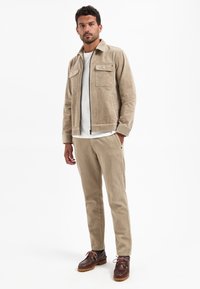 Beige corduroy jas met twee borstzakken, gecombineerd met bijpassende broeken. Bruine bootschoenen met leren accenten maken de outfit af.