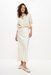 Blusa di raso crema con maniche lunghe, annodata in vita, abbinata a una gonna midi di raso coordinata e scarpe beige con dettagli a fiocco.