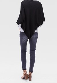 Poncho noir léger avec une coupe large et un ourlet pointu, porté par-dessus une chemise grise à carreaux, assorti à un jean foncé et des chaussures marron à talons blocs.