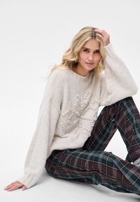 Blondynka siedząca na podłodze, ubrana w kremowy sweter z dzianiny z teksturowanym wzorem śnieżynki oraz zielone i bordowe spodnie w kratę.