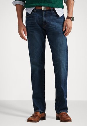 Temno denim hlače s ravnim krojem, z rahlimi madeži in kontrastnim šivanjem. Ujemajo se z rjavimi usnjenimi čevlji in zelenim pletenim puloverjem.