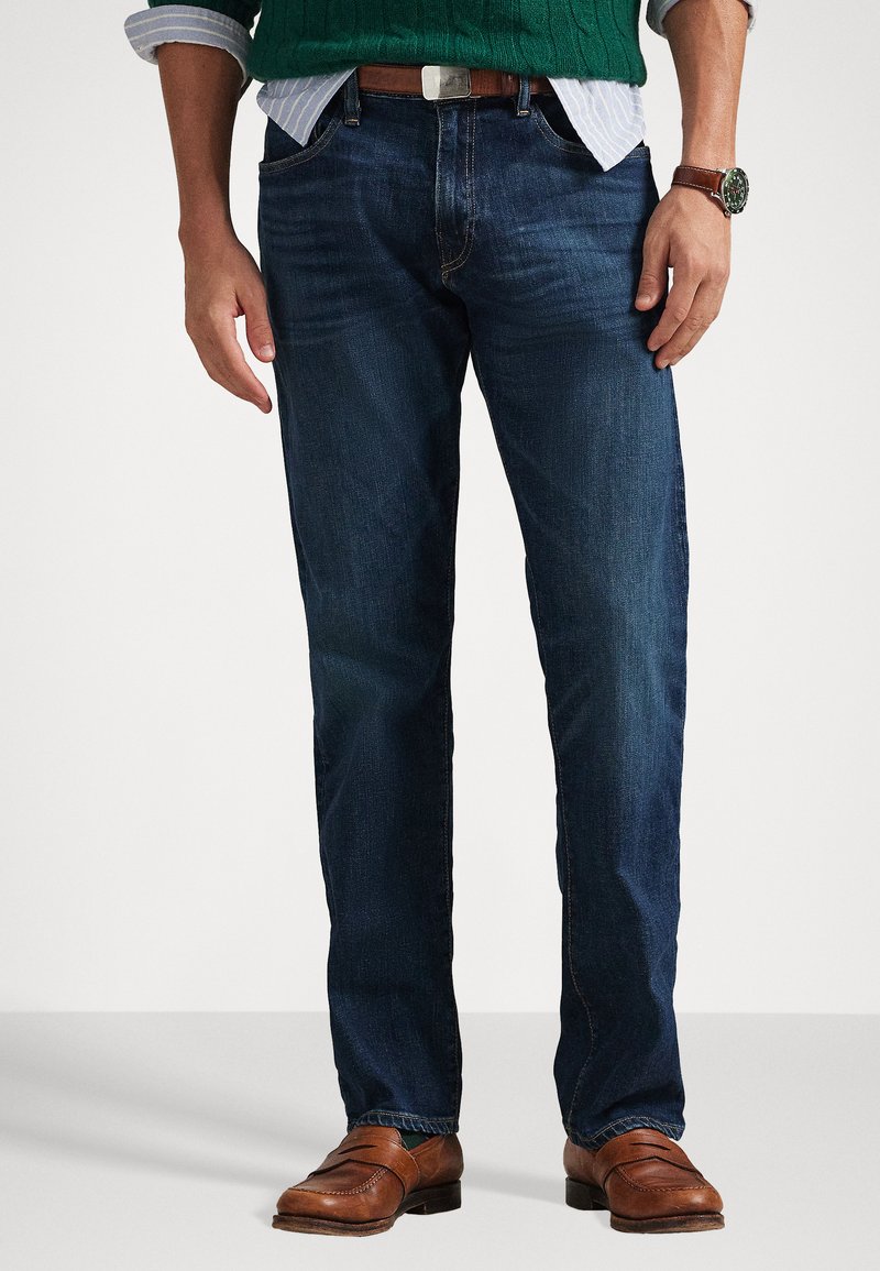 Dark denim jeans with a straight leg fit, subtle fading, and contrast stitching. Paired with brown leather shoes and a green knitted sweater.
