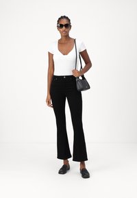 Balta v veida pieguļoša blūze, melni plati džinsi, melni mokasīni un saulesbrilles. Modelis tur mazs melns rokassomu. Minimalistisks dizains.