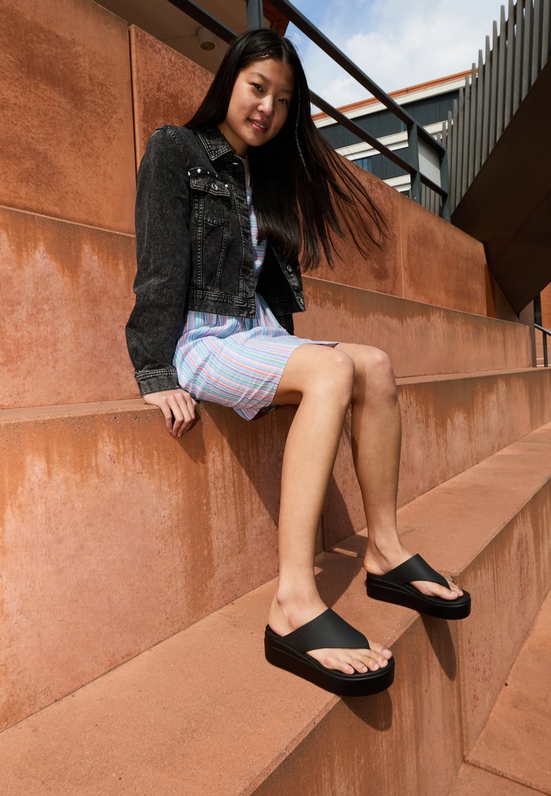 Sandali platform neri con una larga fascia e suola testurizzata, indossati da una persona seduta su gradini in terracotta, che indossa un vestito a quadri e una giacca di denim.