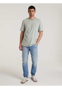 Camiseta verde a rayas con cuello redondo y mangas cortas, combinada con vaqueros azules claros y zapatillas gris con cordones blancos.