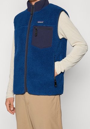 Homme portant une veste polaire bleue avec des poches zippées par-dessus une chemise beige à manches longues et un pantalon marron clair, main dans la poche sur fond gris.