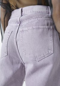 Gros plan d'une personne portant un jean en denim violet clair à taille haute avec des poches arrière et une étiquette de marque visible sur la ceinture.