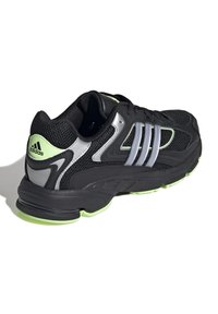 Svart atletisk sko med nätöverdel, silverdetaljer, limegrön sula och en distinkt tre-stripes-design. Dämpad sula för stöd.