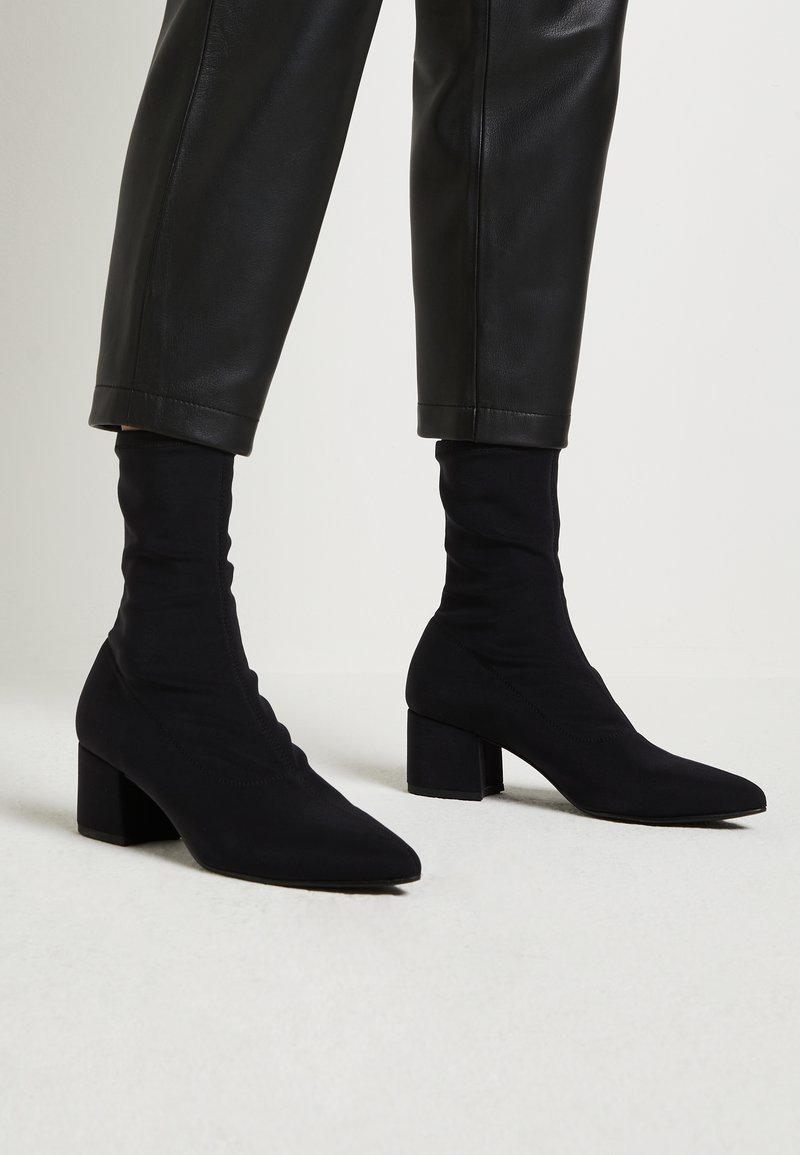 Bottines noires à talons blocs portées avec un pantalon en cuir noir raccourci, présentées sur un fond clair uni.