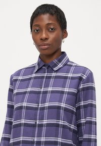 Joven mujer negra con el cabello corto, usando pendientes de aro dorados y una camisa a cuadros morada de botones, mirando con confianza a la cámara.