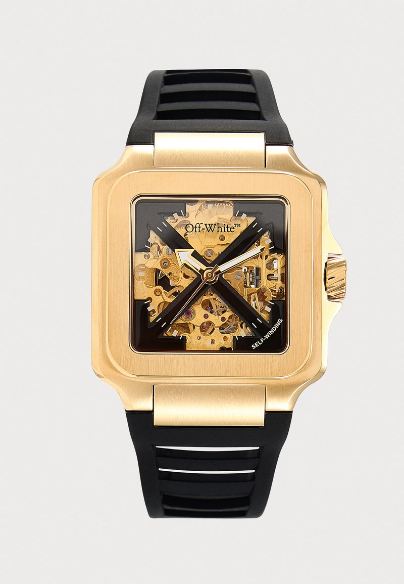 Montre carrée en or avec engrenages mécaniques visibles, aiguilles noires, design noir "X" et bracelet en caoutchouc noir segmenté.