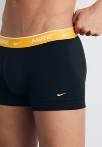 Crne Nike bokserice s žutom elastičnom trakom koja sadrži naziv branda i logo. Glatka tekstura, prilagođeni dizajn i dužina do sredine bedra.
