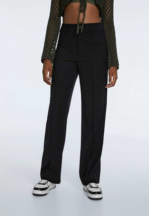 Zwarte high-waisted tailored broek met een rechte pasvorm, voorzien van plooien, zakken en een gladde textuur, gecombineerd met zwart-witte sneakers.