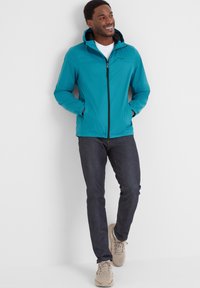 Teal lightweight jacket with a black zip, attached hood, and elastic cuffs, paired with dark jeans and beige sneakers.