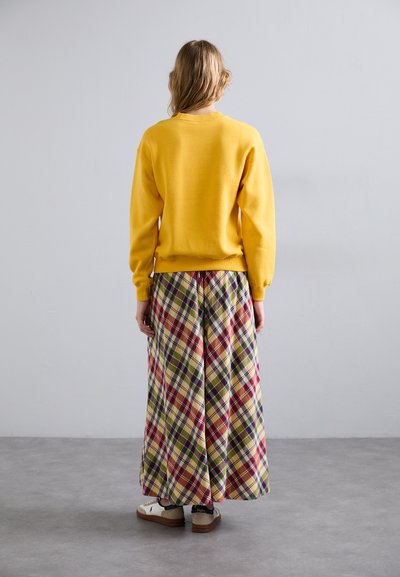 Sudadera amarilla con puños acanalados, combinada con una falda larga de cuadros en tonos rojos, verdes y amarillos. Zapatos blancos con suelas marrones.