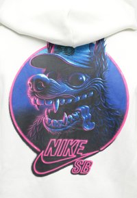 Sweatshirt branco com uma ilustração de um skull de cão a usar um boné azul, com detalhes em rosa e azul, e o texto "NIKE SB" abaixo.