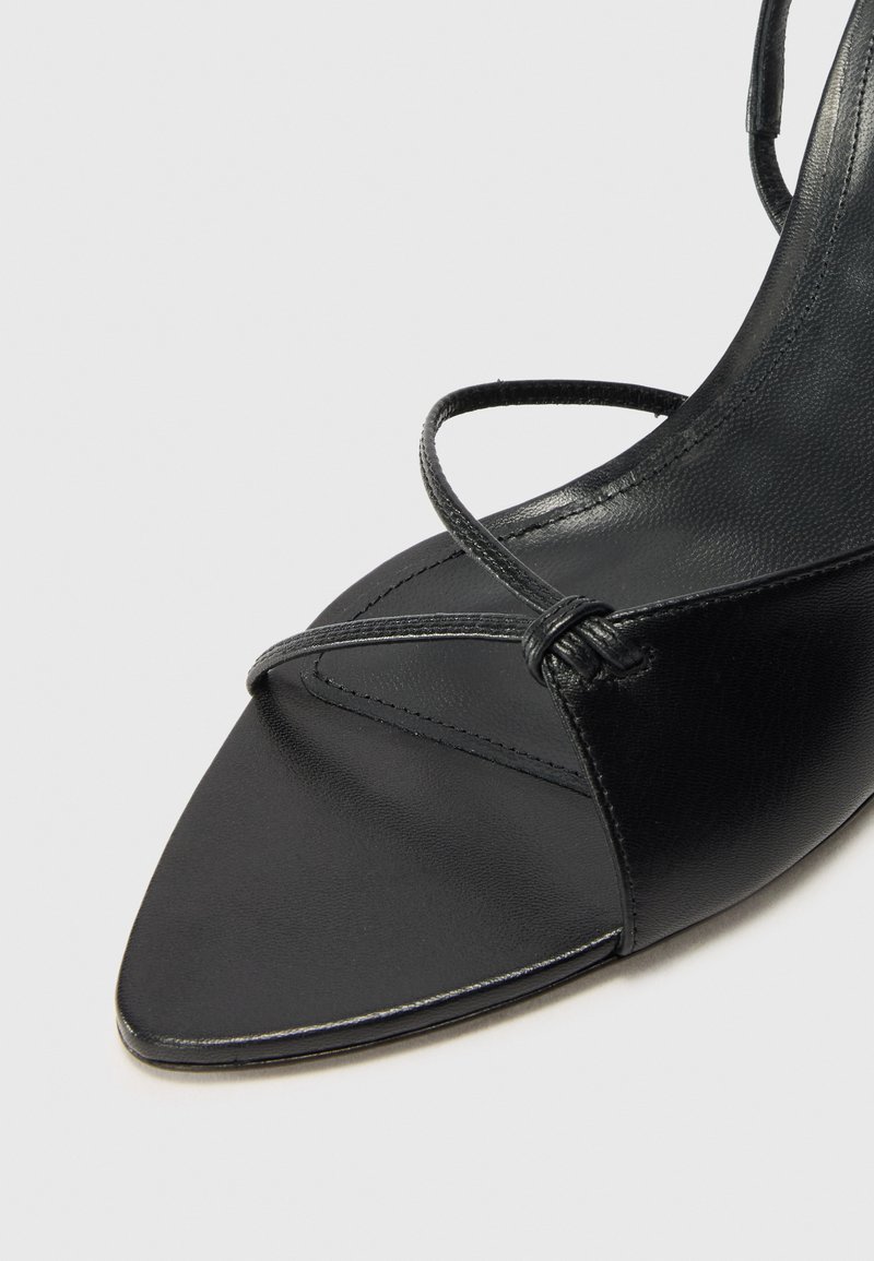 Sandale en cuir noir avec un bout pointu, dotée de fines lanières et d'une texture lisse. Le design met en avant le minimalisme et l'élégance.