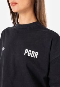 Melna kokvilnas sporta krekls ar ribotu apkakli, īsām piedurknēm un baltu drukātu tekstu "PGDR" priekšpusē. Mīksta tekstūra, brīvs piegriezums.