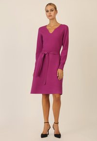 Fuchsia Strickkleid mit V-Ausschnitt, langen Ärmeln und einem gebundenen Taillendetail. Der Saum ist leicht ausgestellt und es wird mit schwarzen Absatzschuhen kombiniert.