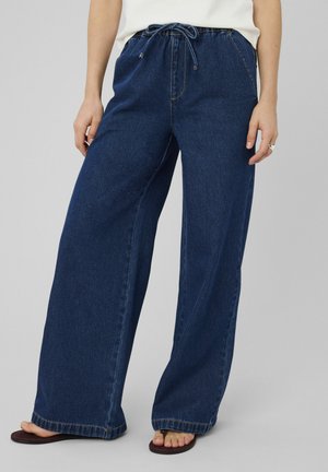 Personne portant un pantalon large en denim bleu foncé avec une taille à cordon, un haut blanc, debout pieds nus sur des sandales sur un fond gris.