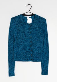 Modrý cardigan s texturovaným vzorem, který má černé čtverce, kulatý výstřih a zapínání na přední knoflíky. Visí na černém háčku.