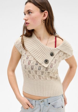 Ung kvinde med langt brunt hår iført en beige strikket ærmeløs top med to store knapper og en synlig rød bh-strop.