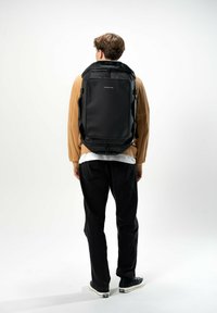 Zaino nero dal design elegante, realizzato in materiale resistente. Presenta spallacci imbottiti, un manico superiore e un branding minimalista sulla parte anteriore.