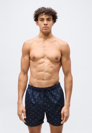 Jonge man met krullend haar die donkerblauwe zwemshort met patroon draagt, staand tegen een effen lichte achtergrond.