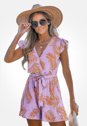 Kobieta ubrana w lawendowy kombinezon z pomarańczowym nadrukiem liści, brązowy słomkowy kapelusz z szerokim rondem, okulary przeciwsłoneczne i trzymająca plecioną torbę.