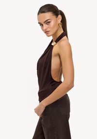 Haut marron à col licou avec un dos ouvert, présentant un tissu drapé et un accent doré au niveau du décolleté, associé à un pantalon taille haute marron foncé.