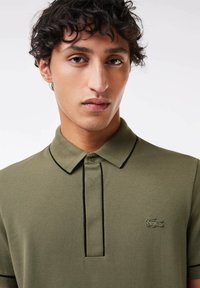 Polo de color verde oliva con tejido texturizado, que presenta un sutil ribete negro y un logotipo bordado en el lado izquierdo del pecho.