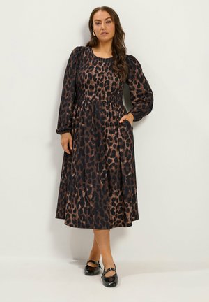 Femme portant une robe à manches longues, longueur mi-mollet, imprimé léopard, avec des chaussures Mary Jane noires, debout devant un fond clair uni.