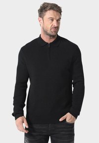 Svart strikket genser med glidelås i kragen, lange ermer og tettsittende design. Stoffet ser mykt ut med en jevn tekstur.