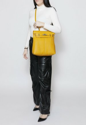 Donna che indossa un dolcevita bianco e pantaloni in pelle nera, tiene una borsa a tracolla gialla in pelle con chiusura argentata, in piedi davanti a uno sfondo neutro.