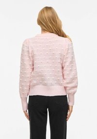 Femme aux longs cheveux blonds portant un pull tricoté rose pâle avec des pois texturés et un pantalon noir, vue de dos sur un fond blanc.
