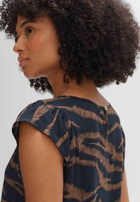 Marineblaue Bluse mit braunem, abstraktem Muster, kurzen Flatterärmeln und Knopfverschluss am Rücken. Glatter Stoff mit dezentem Schimmer.