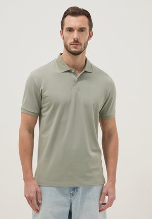 Homme portant un polo vert sauge à manches courtes et un jean bleu clair, debout devant un fond clair uni.