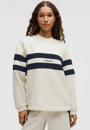 Kobieta ubrana w kremową bluzę z granatowymi poziomymi paskami i logo "lululemon", w zestawie z pasującymi kremowymi spodniami na jednolitym tle.