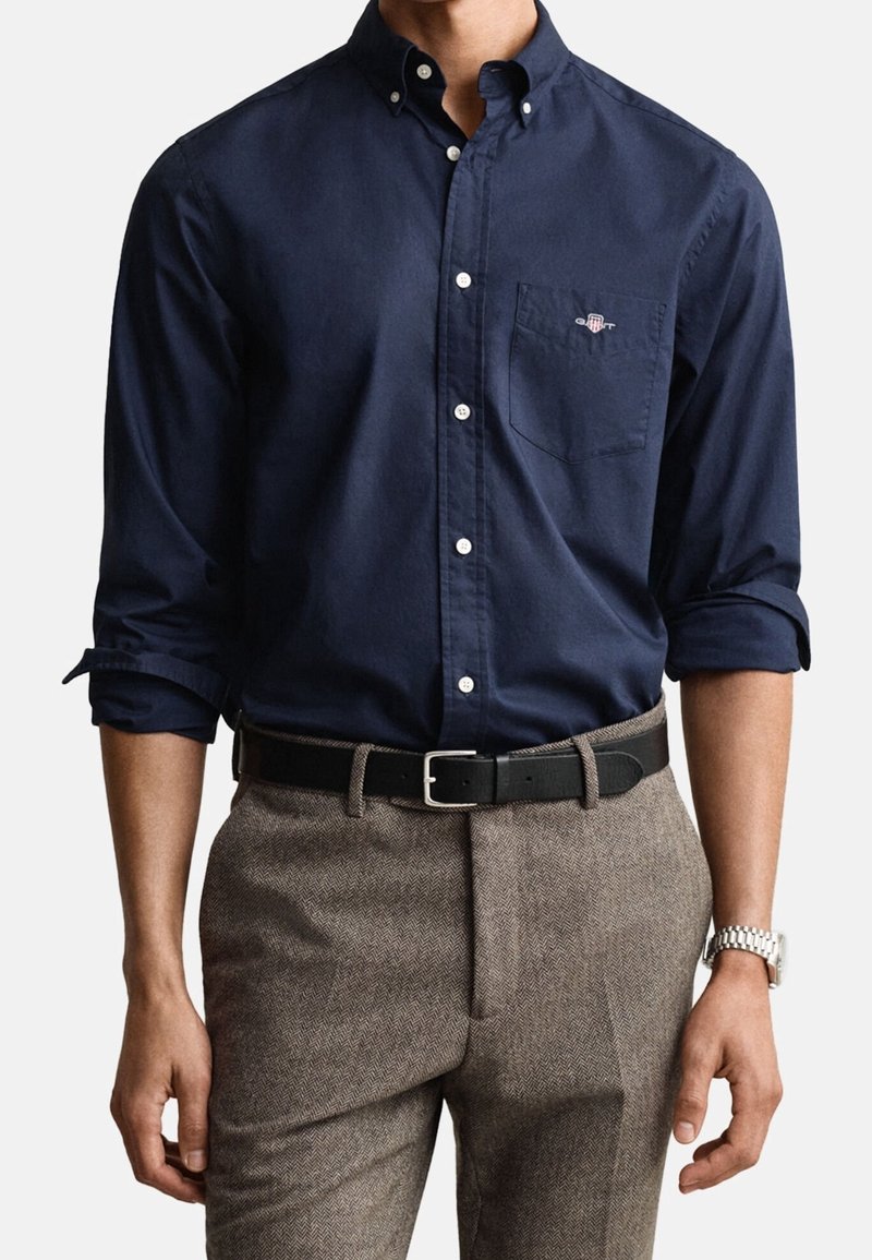 Homme portant une chemise bleu marine boutonnée avec les manches retroussées, un pantalon gris à chevrons, une ceinture noire et une montre-bracelet argentée.