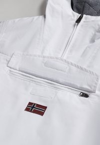 Veste isolée blanche avec une poche frontale zippée, un rabat en Velcro et un petit drapeau norvégien brodé sur la poitrine. Texture douce.