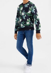 Sudadera negra con estampado floral en verde y blanco, con puños y bajo acanalados, combinada con jeans ajustados azules y zapatillas blancas.