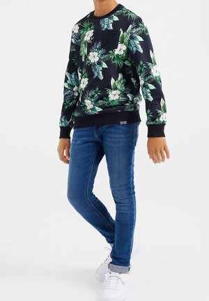Sudadera negra con estampado floral en verde y blanco, con puños y bajo acanalados, combinada con jeans ajustados azules y zapatillas blancas.