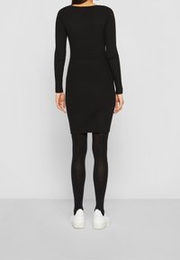Robe noire à côtes avec des manches longues, silhouette ajustée et encolure ronde, assortie à des collants noirs et des baskets blanches.