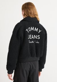 Veste en polaire noire et duveteuse avec "TOMMY JEANS" brodé au dos. Poignets et ceinture côtelés, avec un design de col châle.