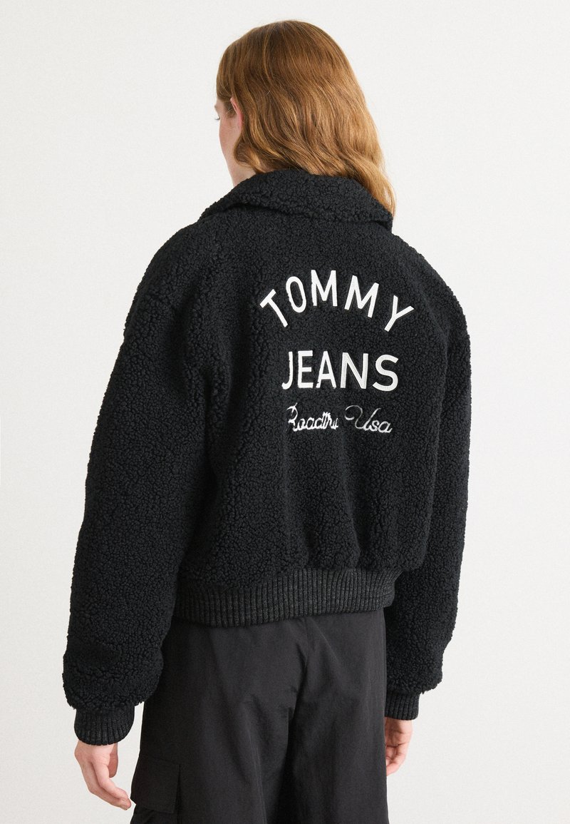 Veste en polaire noire et duveteuse avec "TOMMY JEANS" brodé au dos. Poignets et ceinture côtelés, avec un design de col châle.