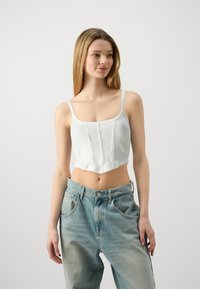 Top court blanc à bretelles fines, avec un côtelé vertical et un décolleté droit, associé à un jean bleu clair taille haute.