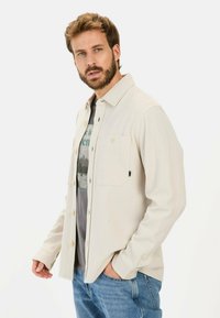 Beige, struktureret skjorte med knapper og en brystlomme, med en lige krave og lange ærmer, båret over en grafisk t-shirt og blå jeans.