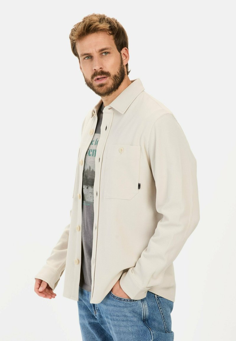Beige, struktureret skjorte med knapper og en brystlomme, med en lige krave og lange ærmer, båret over en grafisk t-shirt og blå jeans.